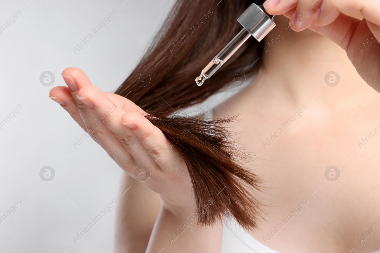 Photo of Woman applying essential oil onto hair on light grey background, closeup