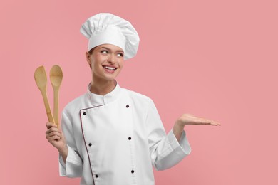 Photo of Happy chef in uniform holding wooden spatula and spoon on pink background, space for text