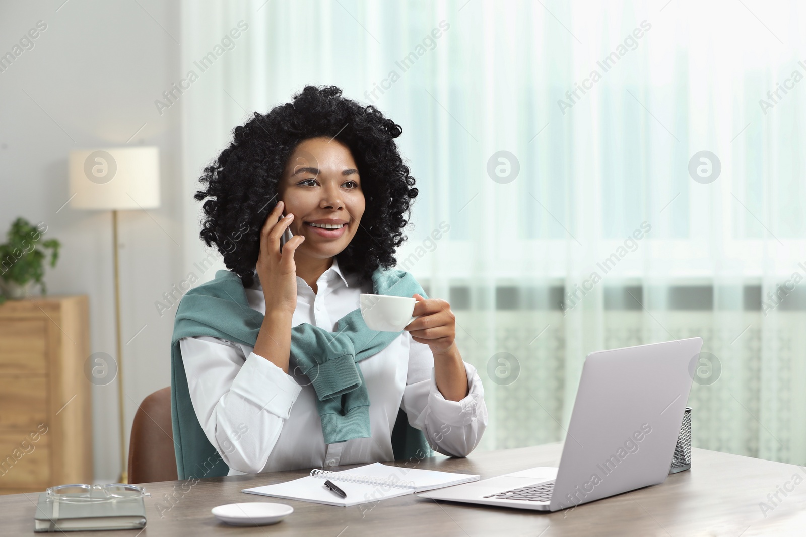 Photo of Happy young woman with cup of drink talking on phone near laptop at wooden desk indoors