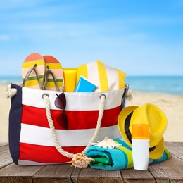 Image of Stylish bag with beach accessories on wooden surface near seashore 