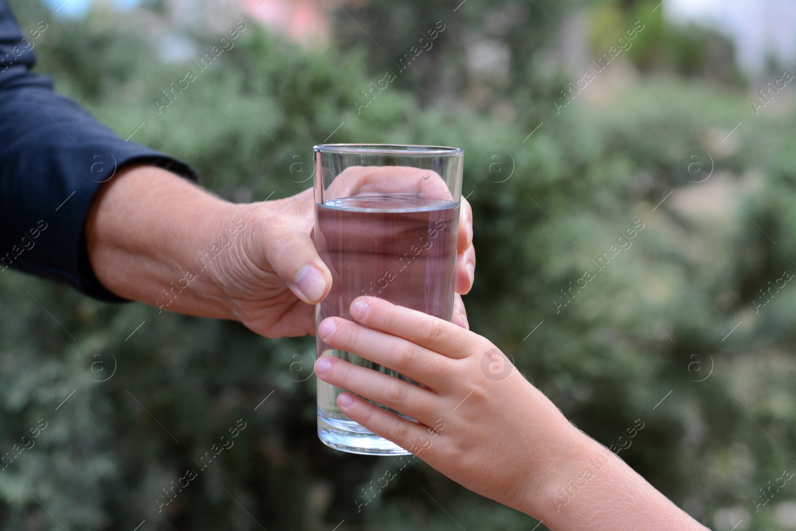 Photo of Child giving glass of water to elderly woman outdoors, closeup