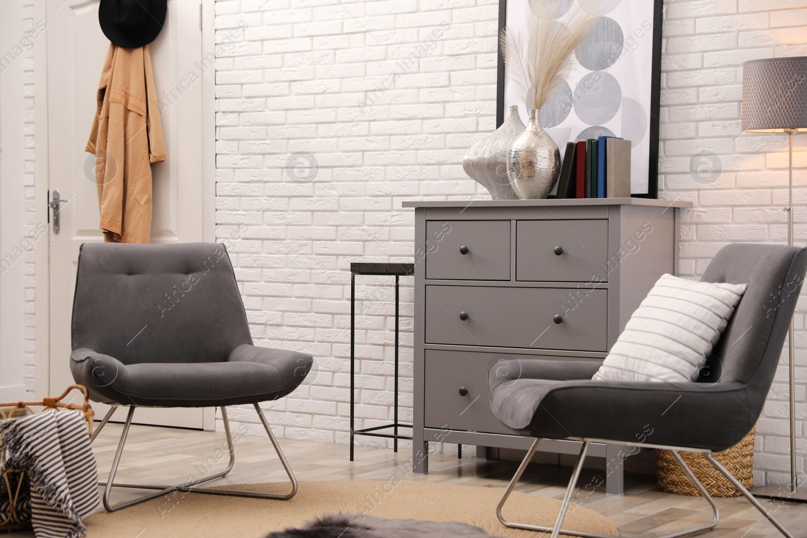 Photo of Stylish room interior with grey chest of drawers and chairs near white brick wall