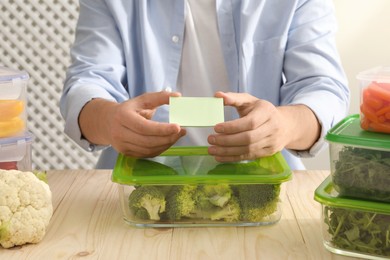 Photo of Man sticking paper note onto container with fresh broccoli at wooden table, closeup. Food storage