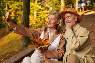 Photo of Affectionate senior couple with dry leaves taking selfie in autumn park