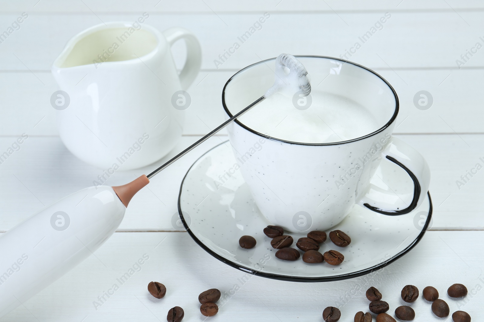 Photo of Mini mixer (milk frother), whipped milk and coffee beans on white wooden table, closeup