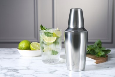 Metal cocktail shaker, delicious mojito, limes and mint on white marble table