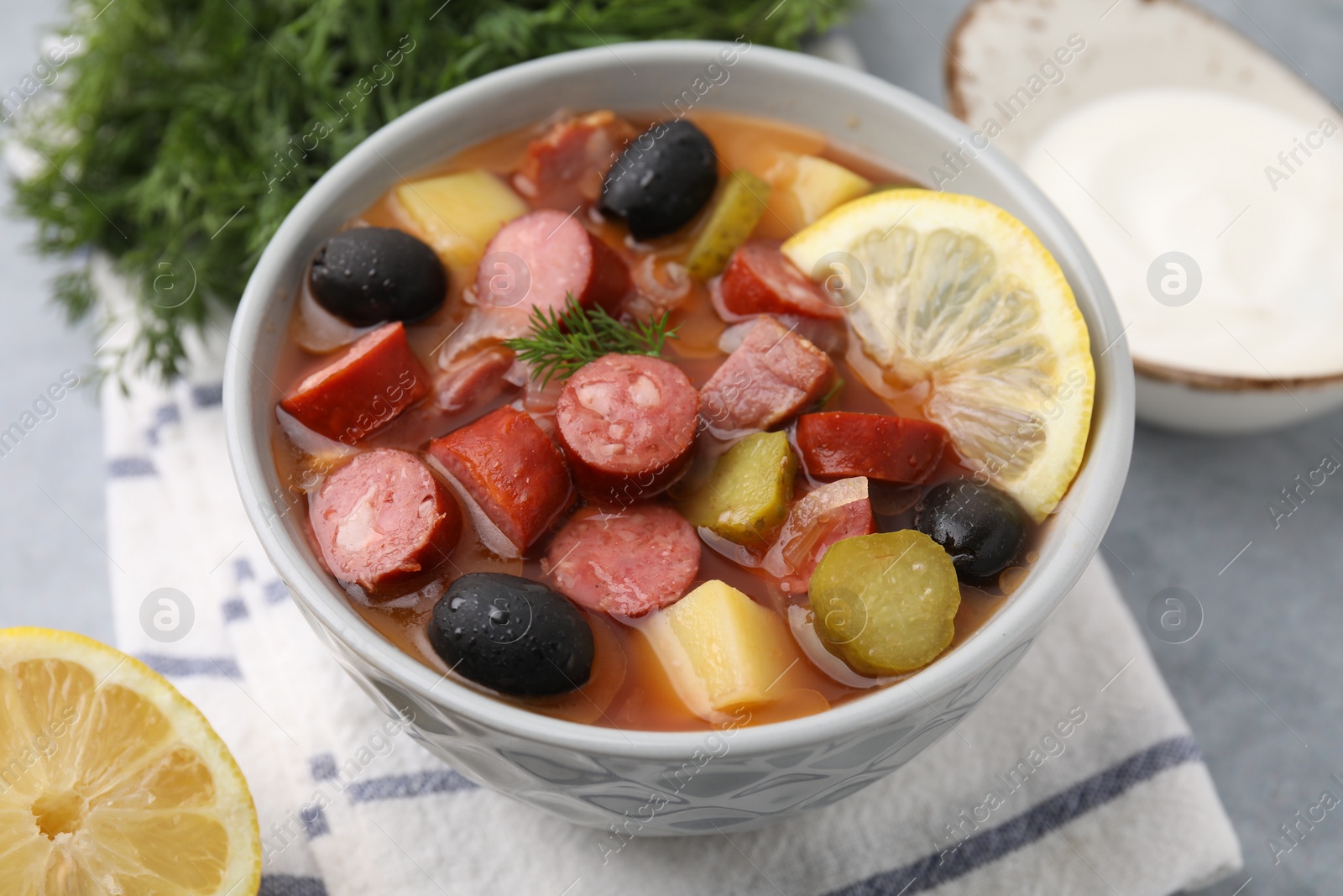 Photo of Meat solyanka soup with thin dry smoked sausages in bowl on grey table, closeup