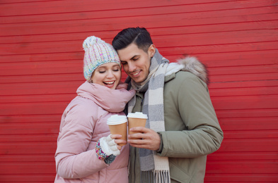 Happy couple in warm clothes with drinks near red wooden wall outdoors. Christmas season