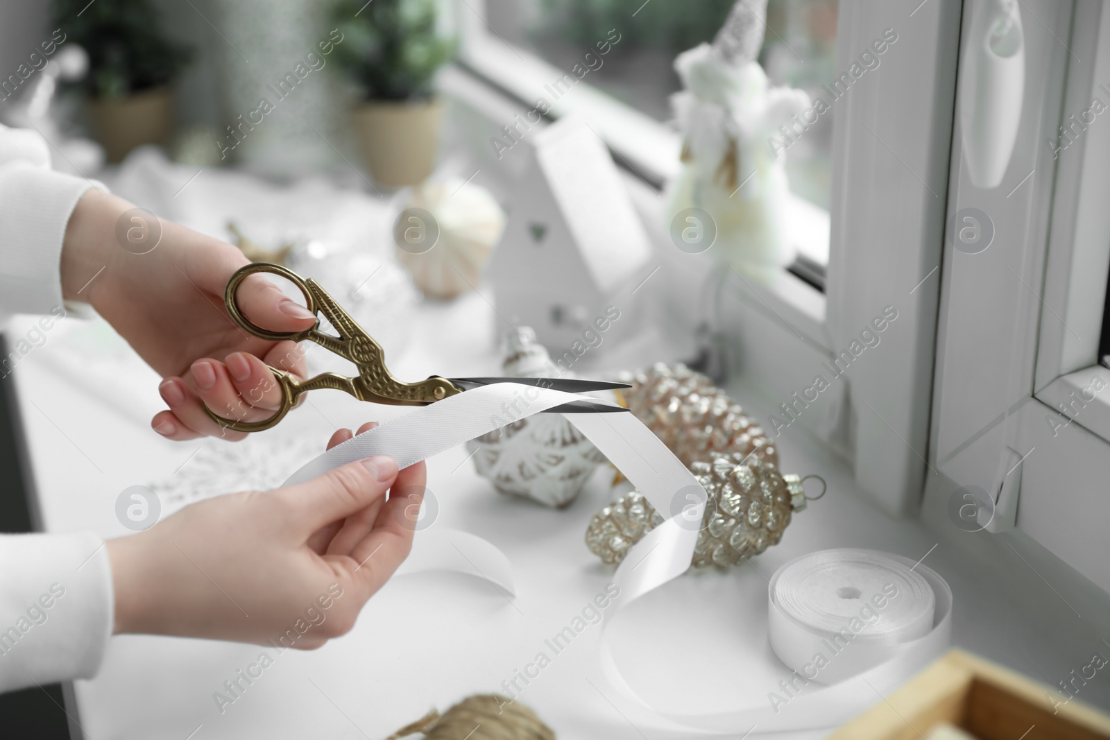 Photo of Woman cutting ribbon near Christmas baubles on window sill indoors, closeup