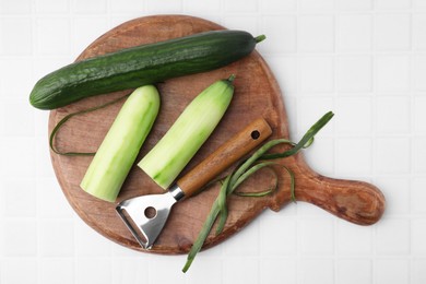 Fresh cucumbers and peeler on white tiled table, top view