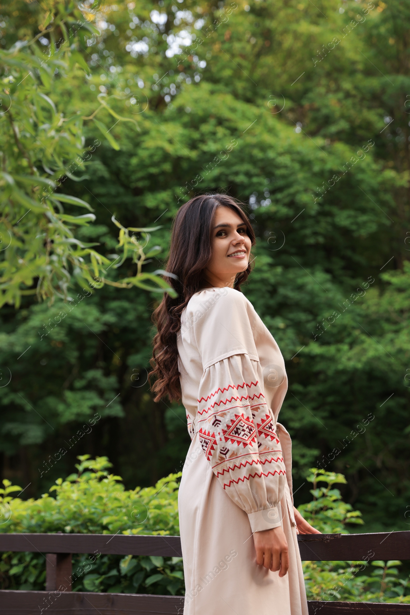 Photo of Beautiful woman wearing embroidered dress near wooden railing in countryside, space for text. Ukrainian national clothes