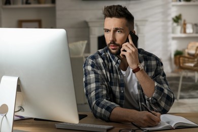Photo of Man talking on phone while working with computer at table in home office