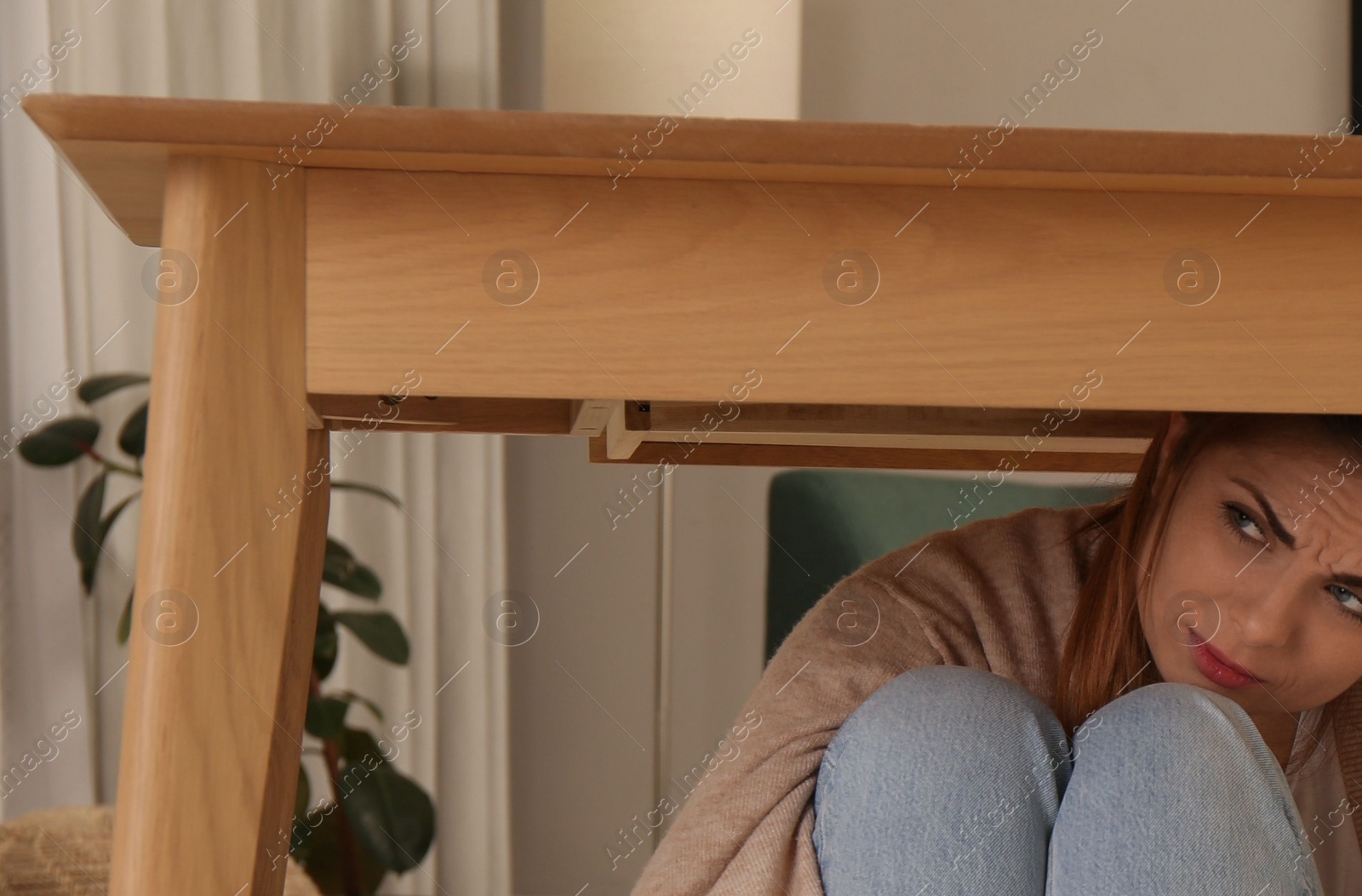 Photo of Scared mother with her little daughter hiding under table in living room during earthquake