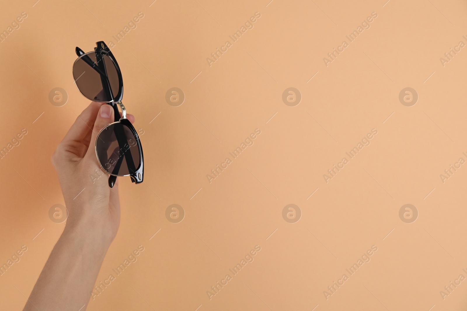 Photo of Woman holding stylish sunglasses on pale orange background, closeup. Space for text