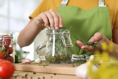 Woman putting herbs into pickling jar at table in kitchen, closeup