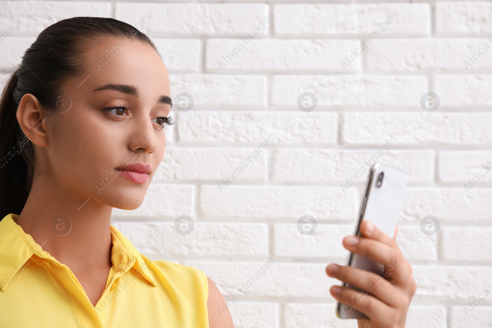 Photo of Young woman unlocking smartphone with facial scanner near white brick wall. Biometric verification