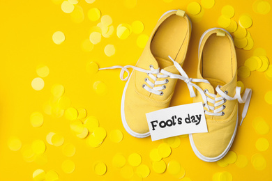 Photo of Shoes tied together, note with phrase FOOL'S DAY and confetti on yellow background, flat lay. Space for text
