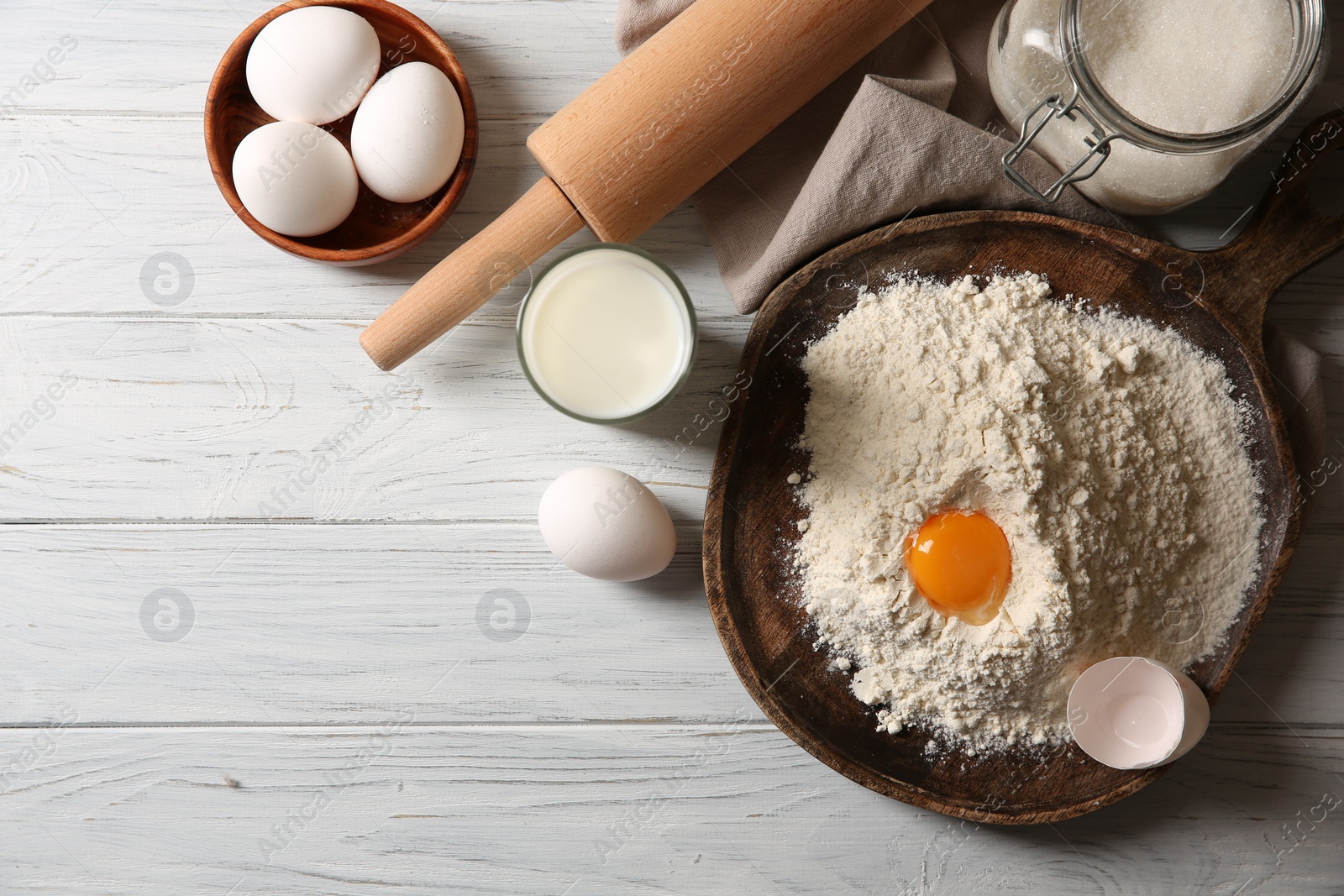 Photo of Pile of flour with yolk and other ingredients for dough on white wooden table, flat lay. Space for text