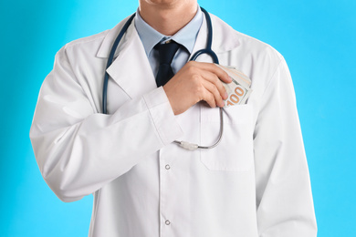 Doctor putting bribe into pocket on light blue background, closeup. Corruption in medicine