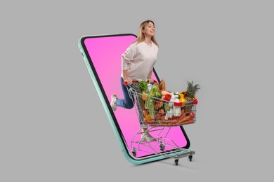 Image of Grocery shopping via internet. Happy woman with shopping cart full of products riding out of huge smartphone on grey background