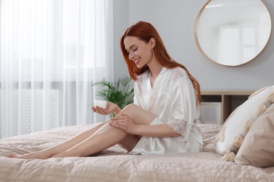 Photo of Beautiful young woman applying body cream onto legs in bedroom