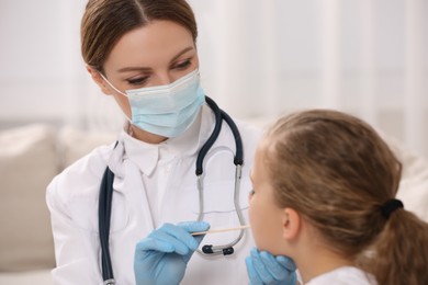Photo of Doctor in medical mask examining girl`s oral cavity with tongue depressor indoors