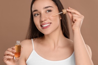 Photo of Beautiful woman applying essential oil onto face on brown background