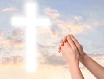 Image of Religion. Christian woman praying against sky with glowing cross, closeup