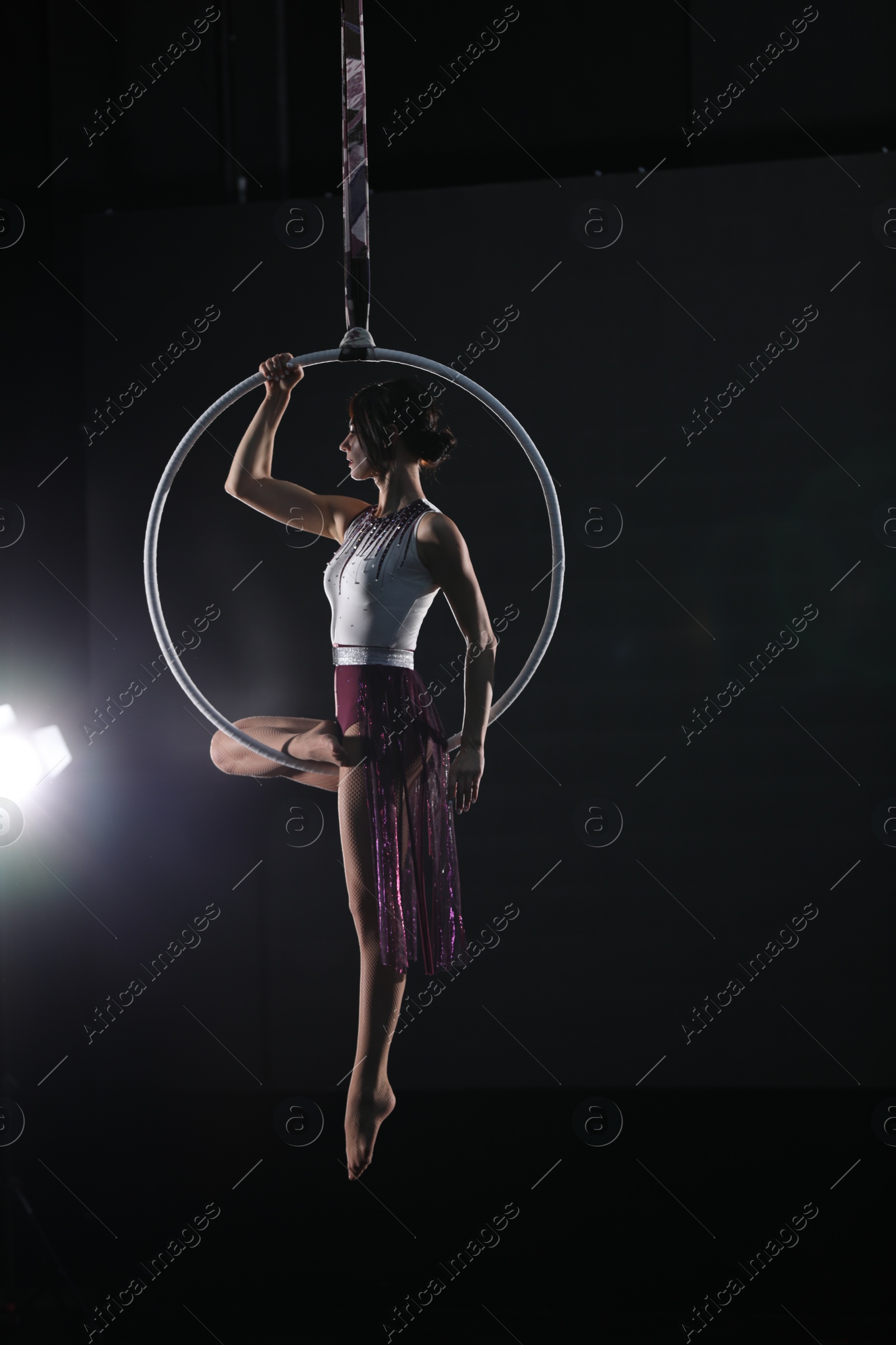 Photo of Young woman performing acrobatic element on aerial ring against dark background