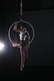Young woman performing acrobatic element on aerial ring against dark background
