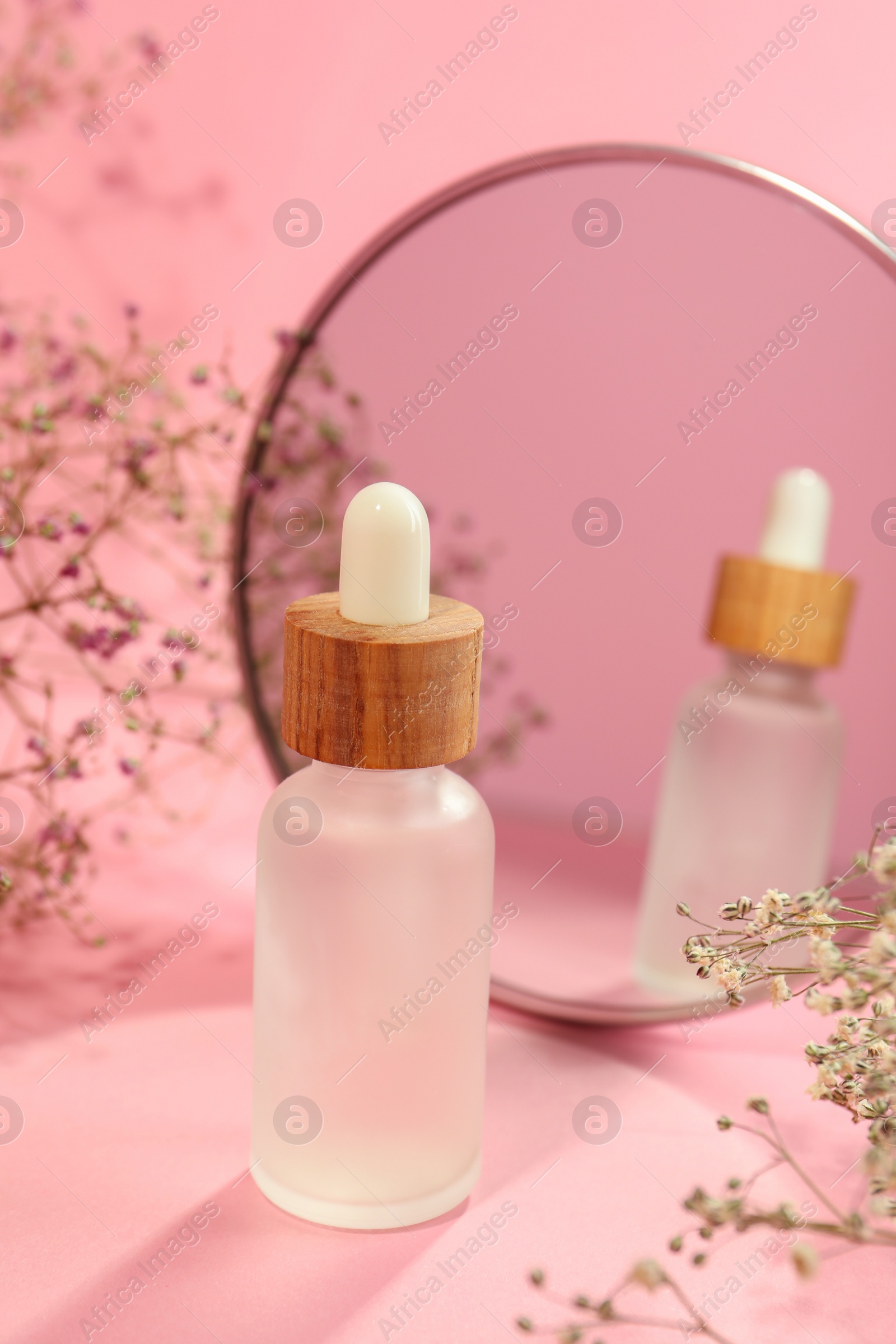 Photo of Bottle of face serum and beautiful flowers near mirror on pink background