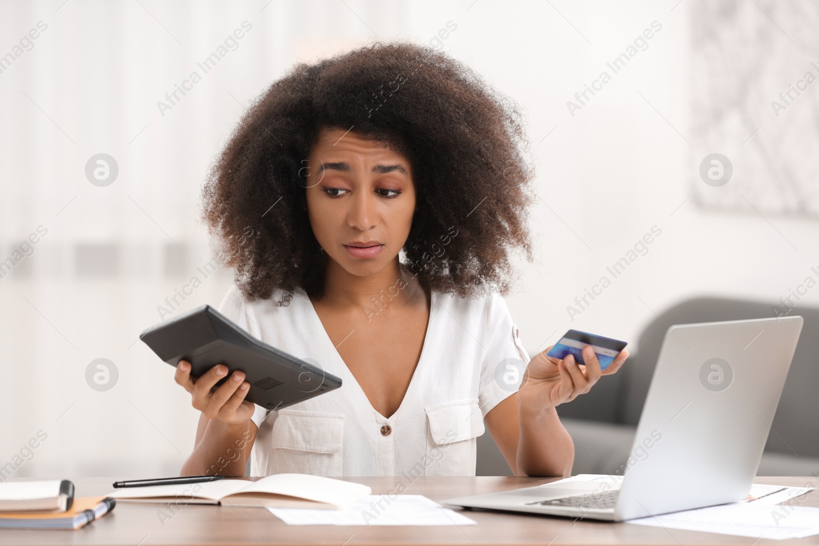 Photo of Confused woman with credit card and calculator planning budget at table indoors. Debt problem