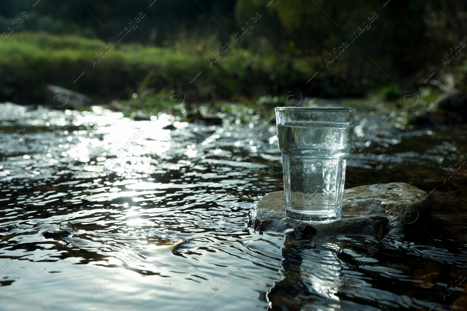 Photo of Glass of fresh water on stone near river outdoors. Space for text