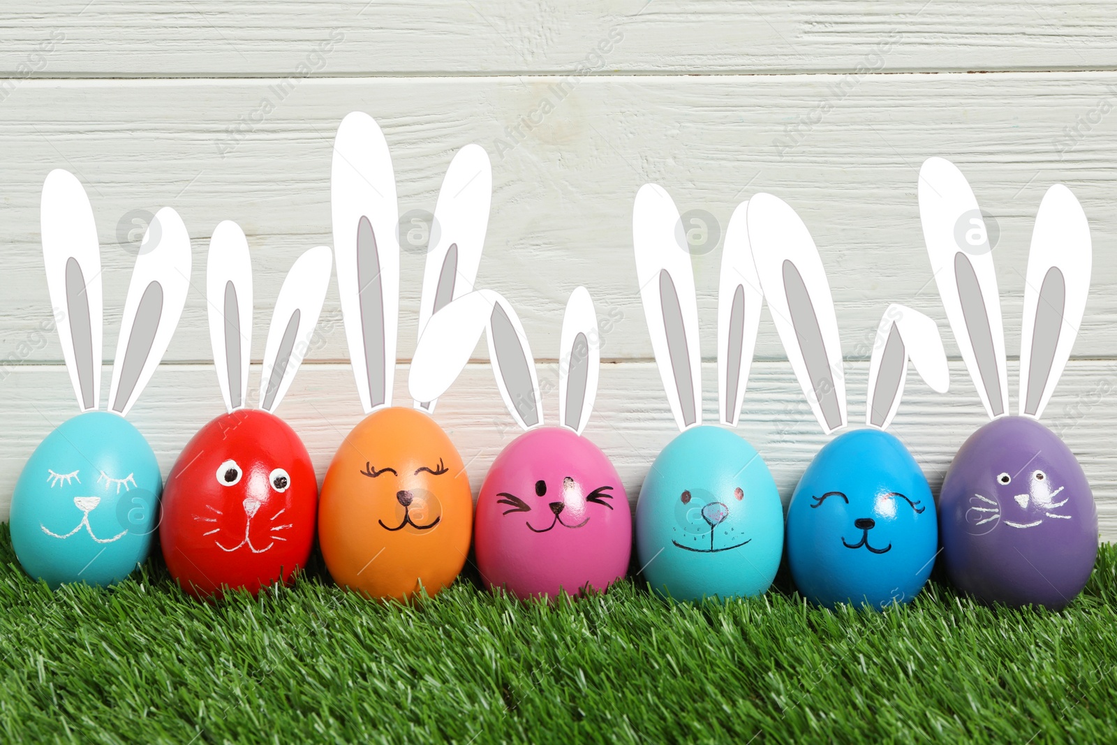 Image of Colorful eggs as Easter bunnies on green grass against white wooden background