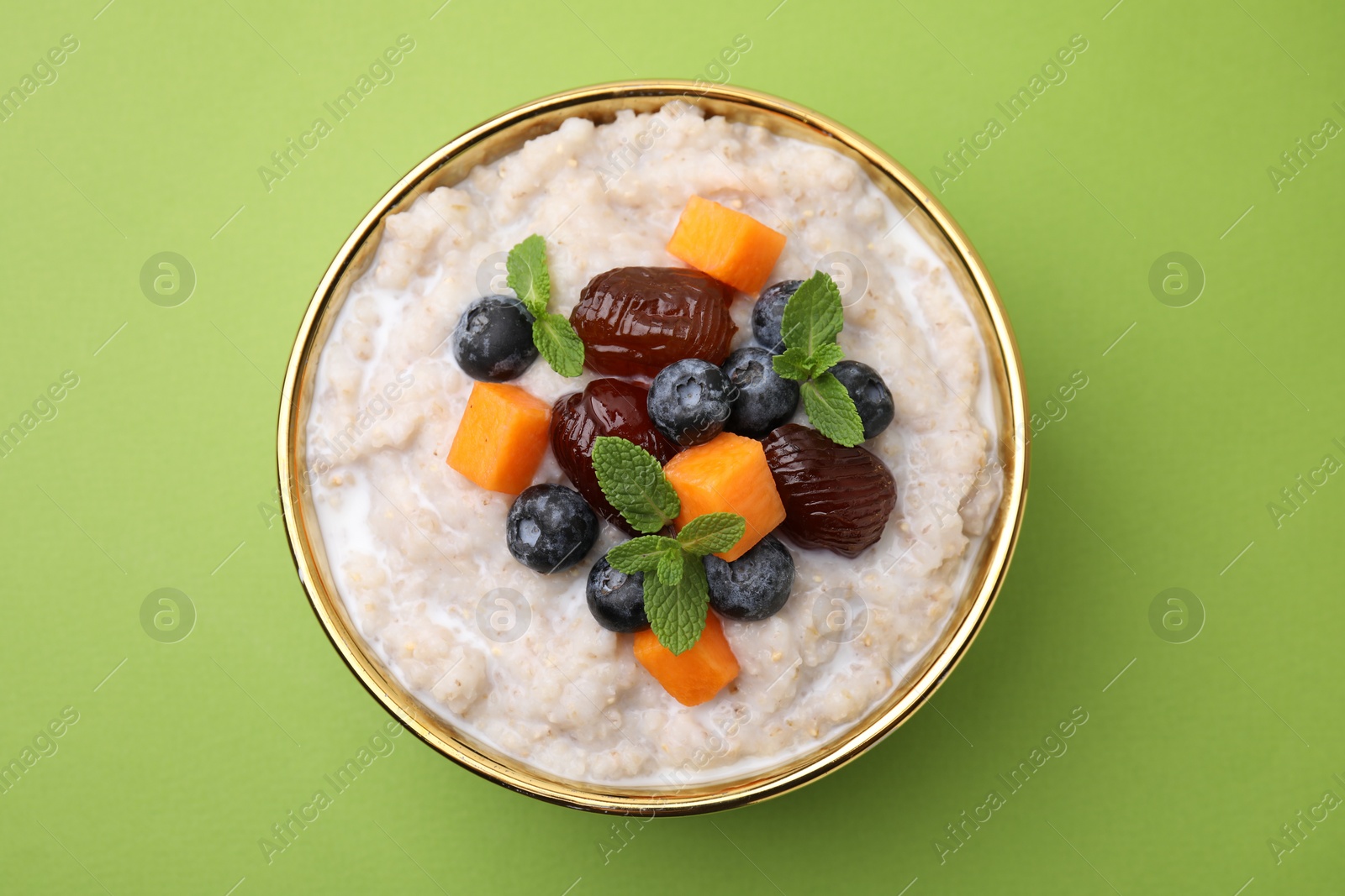 Photo of Delicious barley porridge with blueberries, pumpkin, dates and mint in bowl on green table, top view