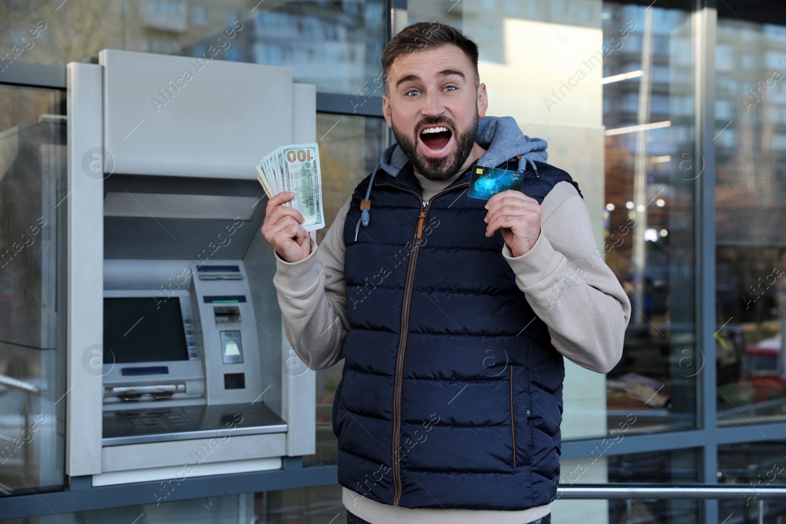 Photo of Excited young man with credit card and money near cash machine outdoors