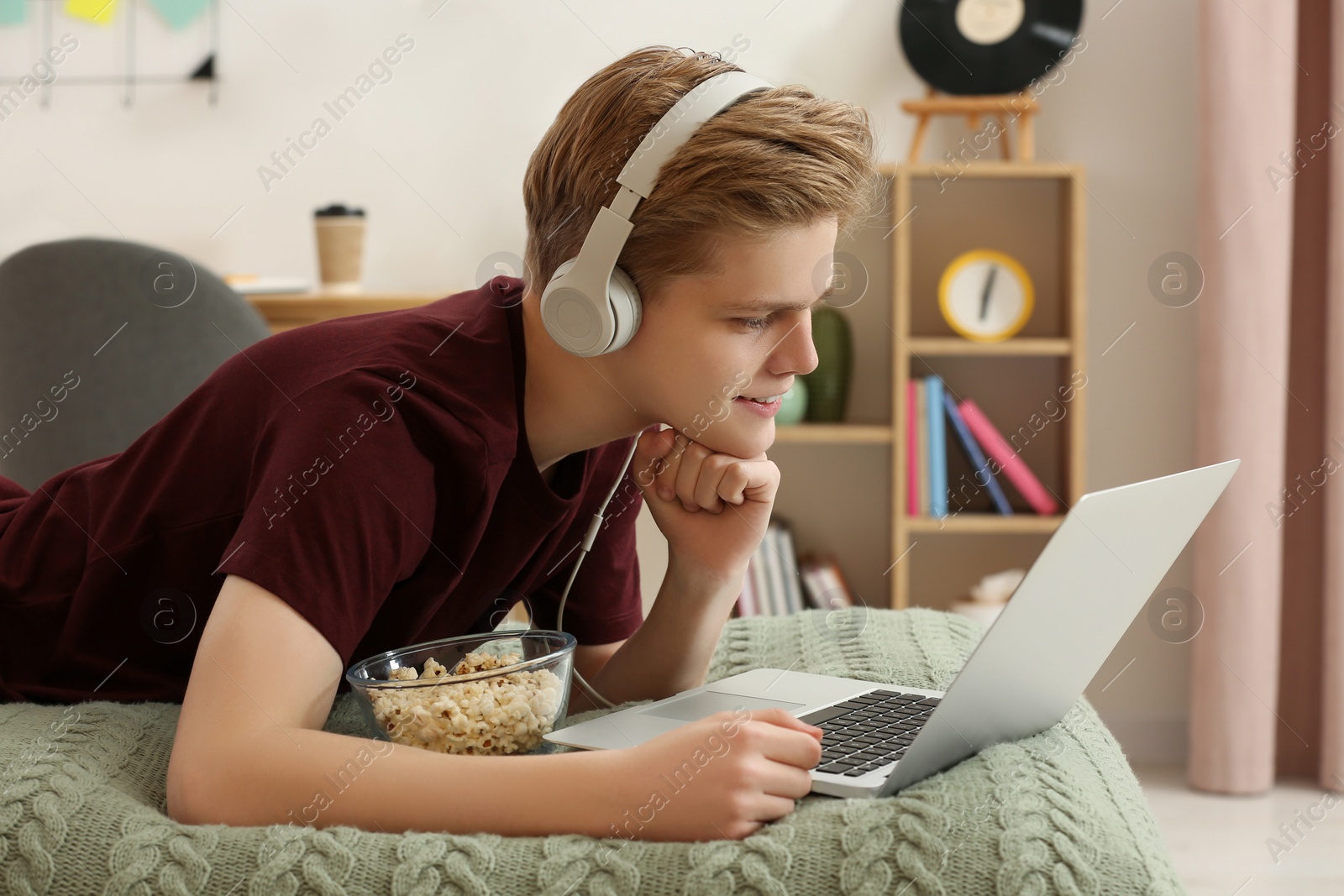 Photo of Teenage boy with headphones and popcorn using laptop on bed at home