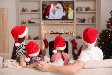 Photo of Family wearing Christmas hats watching TV in cosy room, back view. Winter holidays atmosphere