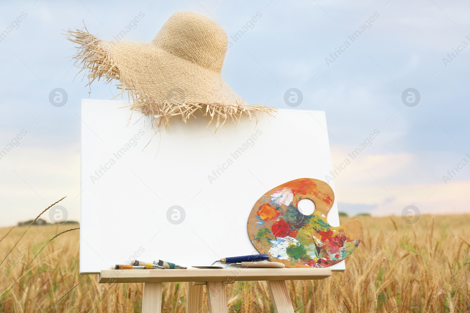 Photo of Wooden easel with blank canvas, painting equipment and hat in field. Space for text