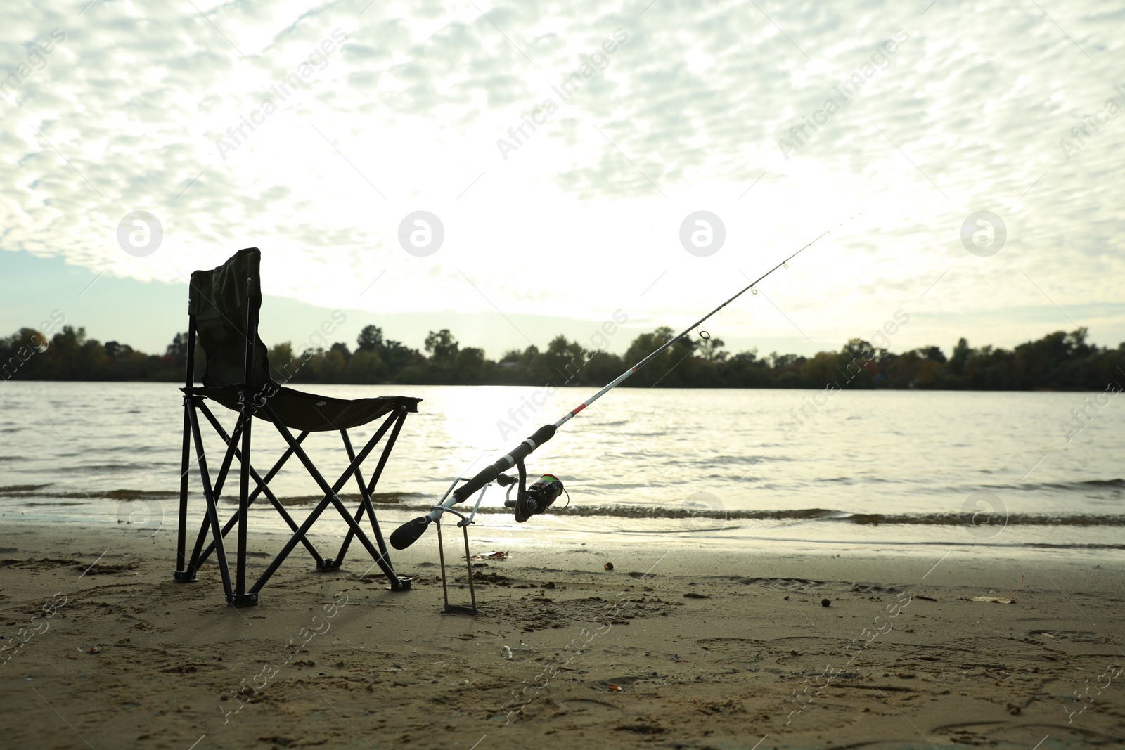 Photo of Folding chair and fishing rod on sand near river, space for text