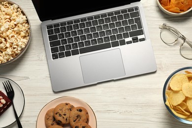 Photo of Bad eating habits at workplace. Laptop, glasses and different snacks on white wooden table, flat lay