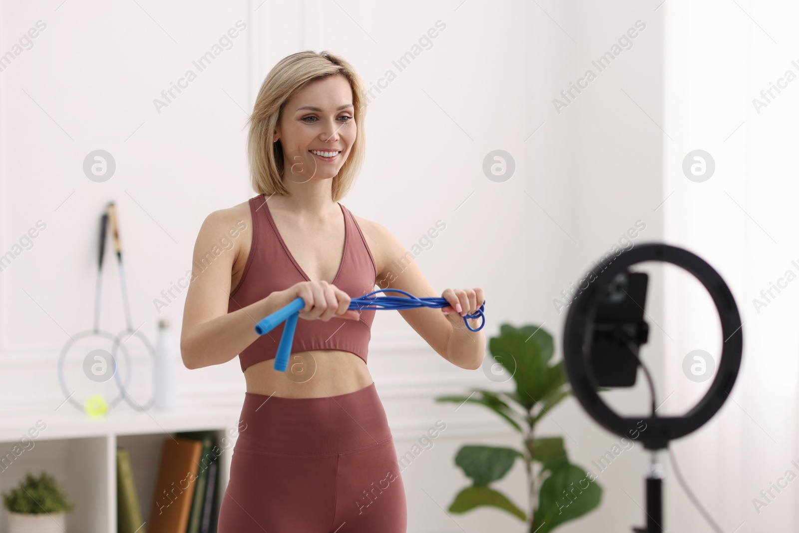 Photo of Smiling sports blogger holding skipping rope while streaming online fitness lesson at home