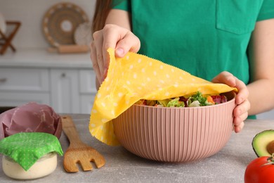 Photo of Woman covering bowl of fresh salad with beeswax food wrap at light grey table in kitchen, closeup