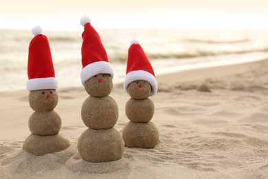 Snowmen made of sand with Santa hats on beach near sea, space for text. Christmas vacation