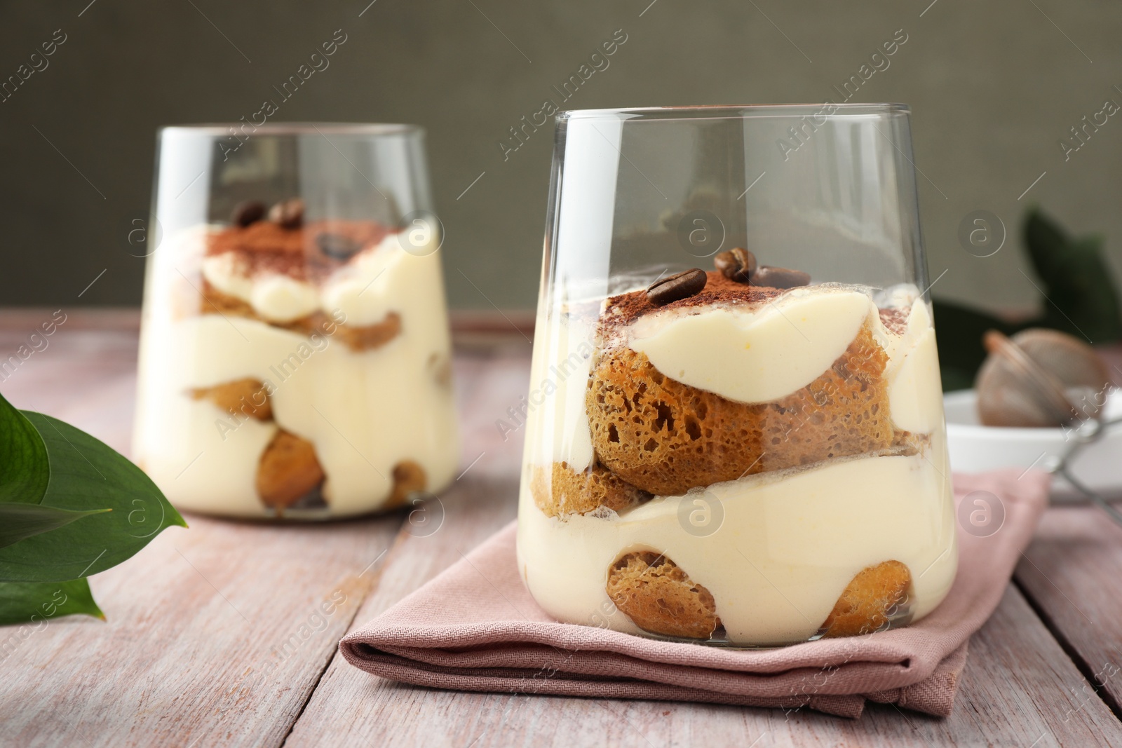 Photo of Delicious tiramisu with coffee beans in glasses and green leaves on wooden table, closeup