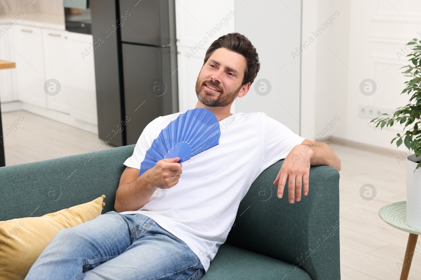 Photo of Bearded man waving blue hand fan to cool himself on sofa at home