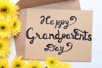 Photo of Beautiful yellow flowers, envelope and card with phrase Happy Grandparents Day on white wooden table, flat lay