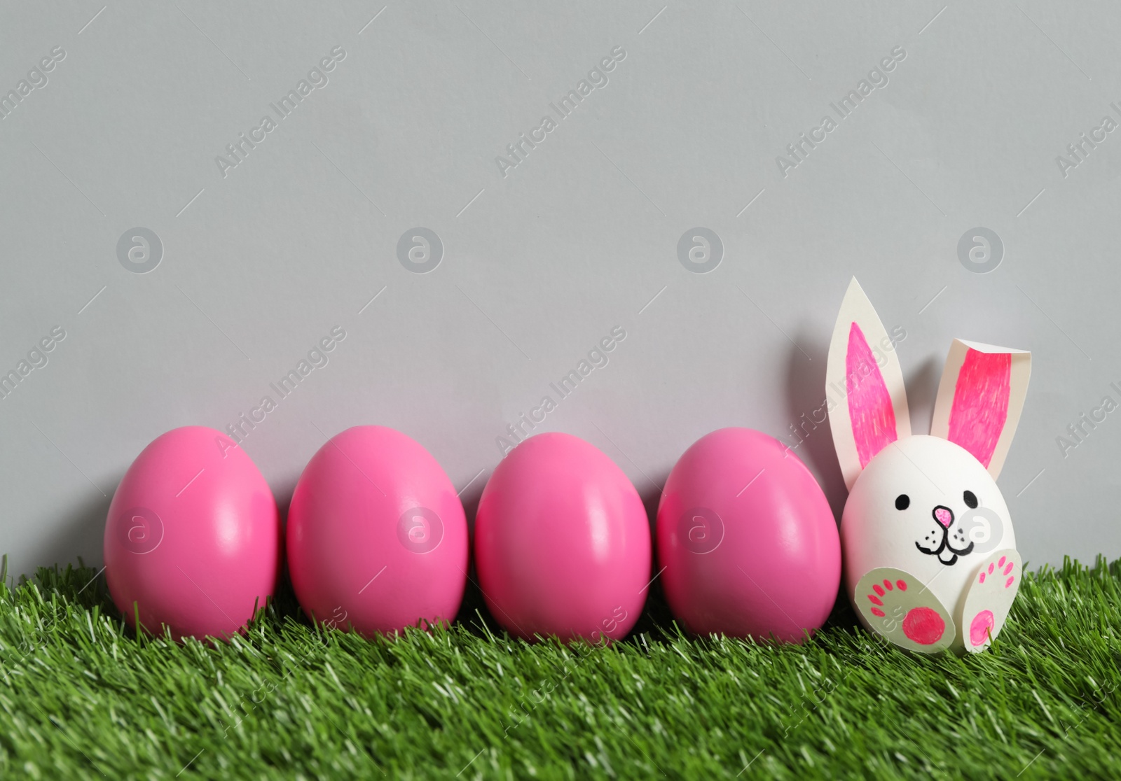 Photo of Bright eggs and white one as Easter bunny on green grass against grey background