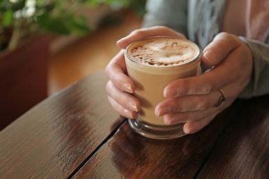 Woman holding cup of aromatic coffee with foam at wooden table