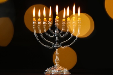 Silver menorah with burning candles against dark background and blurred festive lights. Hanukkah celebration